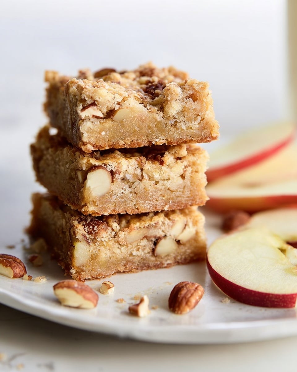 Three thick square bars are stacked on a white plate, each bar showing a crumbly light brown texture with visible chunks of nuts inside. Small pieces of nuts are scattered on the plate around the stack. To the right, thin slices of apple with red edges rest on the white marbled surface. The background is softly blurred, focusing attention on the bars. photo taken with an iphone --ar 4:5 --v 7