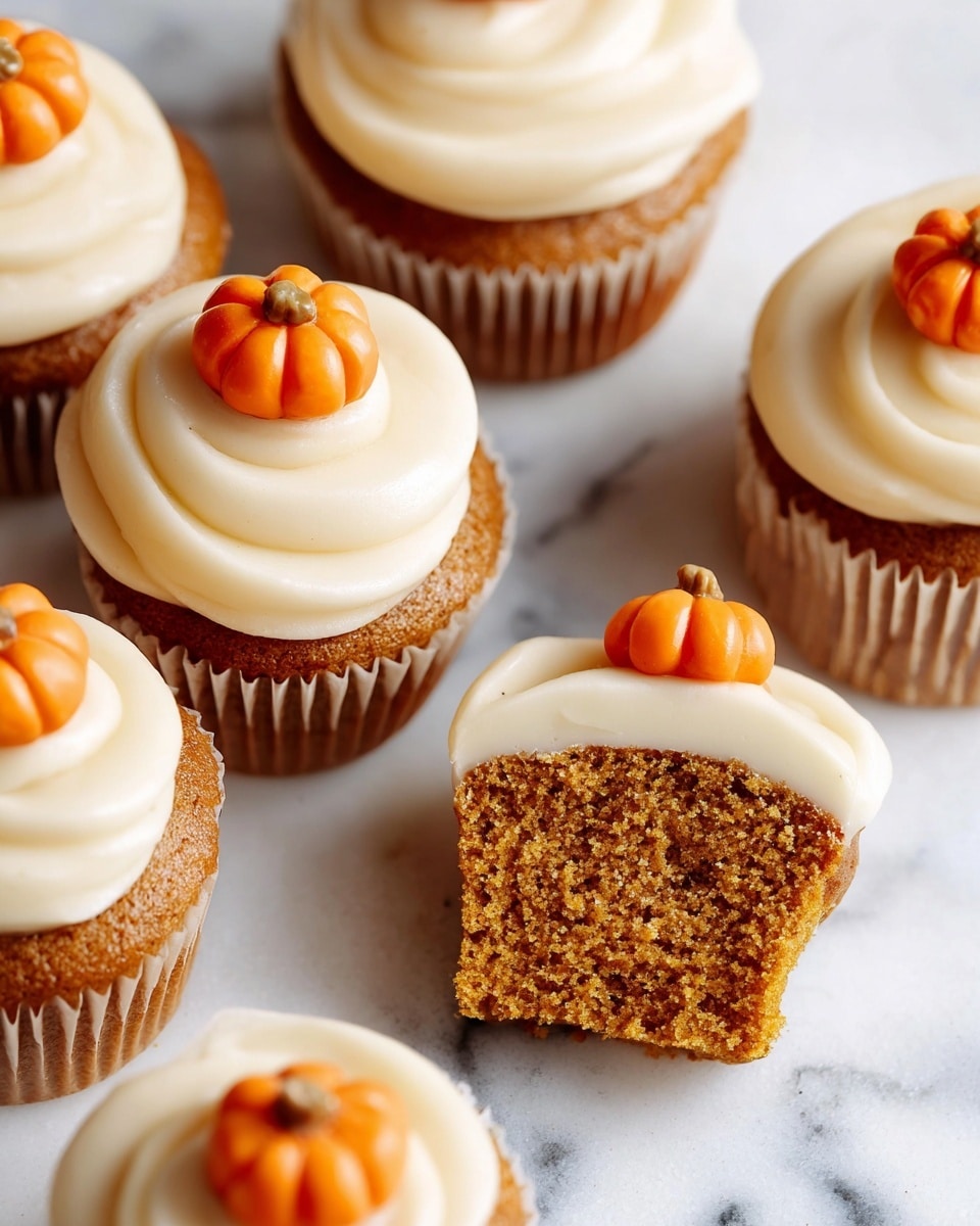The image shows several pumpkin cupcakes arranged on a white marbled surface. Each cupcake has a light brown, moist-looking base with a thick, creamy swirl of pale cream-colored frosting on top. Small, bright orange pumpkin-shaped decorations with detailed ridges and tiny brown stems sit atop each frosting swirl. One cupcake in the center is cut in half to show the soft, crumbly texture inside the brown cake base, topped with the same creamy frosting layer. The colors are warm and inviting, with a contrast between the creamy frosting, the brown cake, and the vivid orange pumpkins. Photo taken with an iphone --ar 4:5 --v 7
