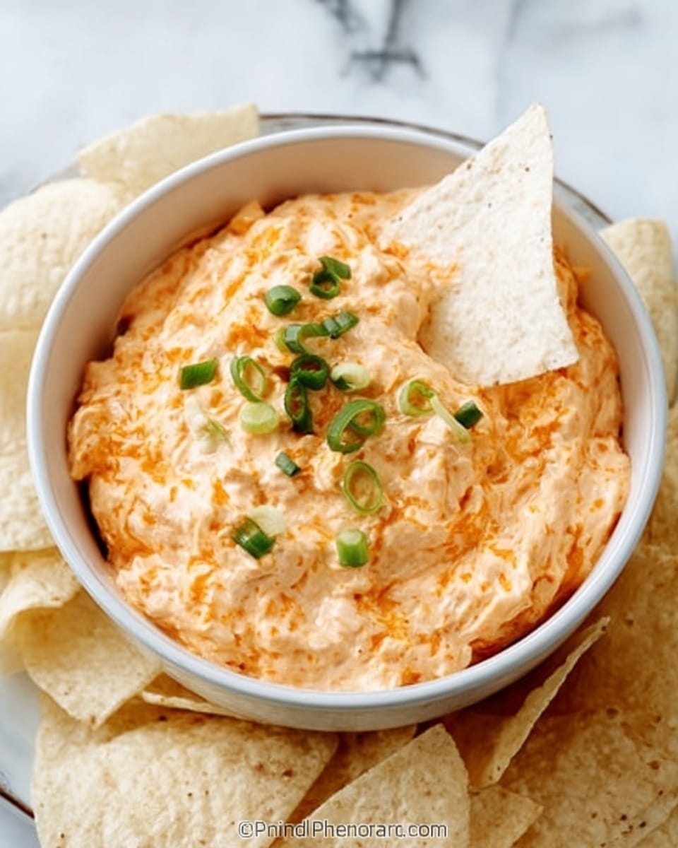 A white bowl filled with a creamy orange dip that has a rough, chunky texture. On top, there are small green onion pieces scattered, and a single triangular white tortilla chip is dipped in the center. The bowl is surrounded by more white tortilla chips placed around it. The scene is set on a white marbled surface. Photo taken with an iphone --ar 4:5 --v 7