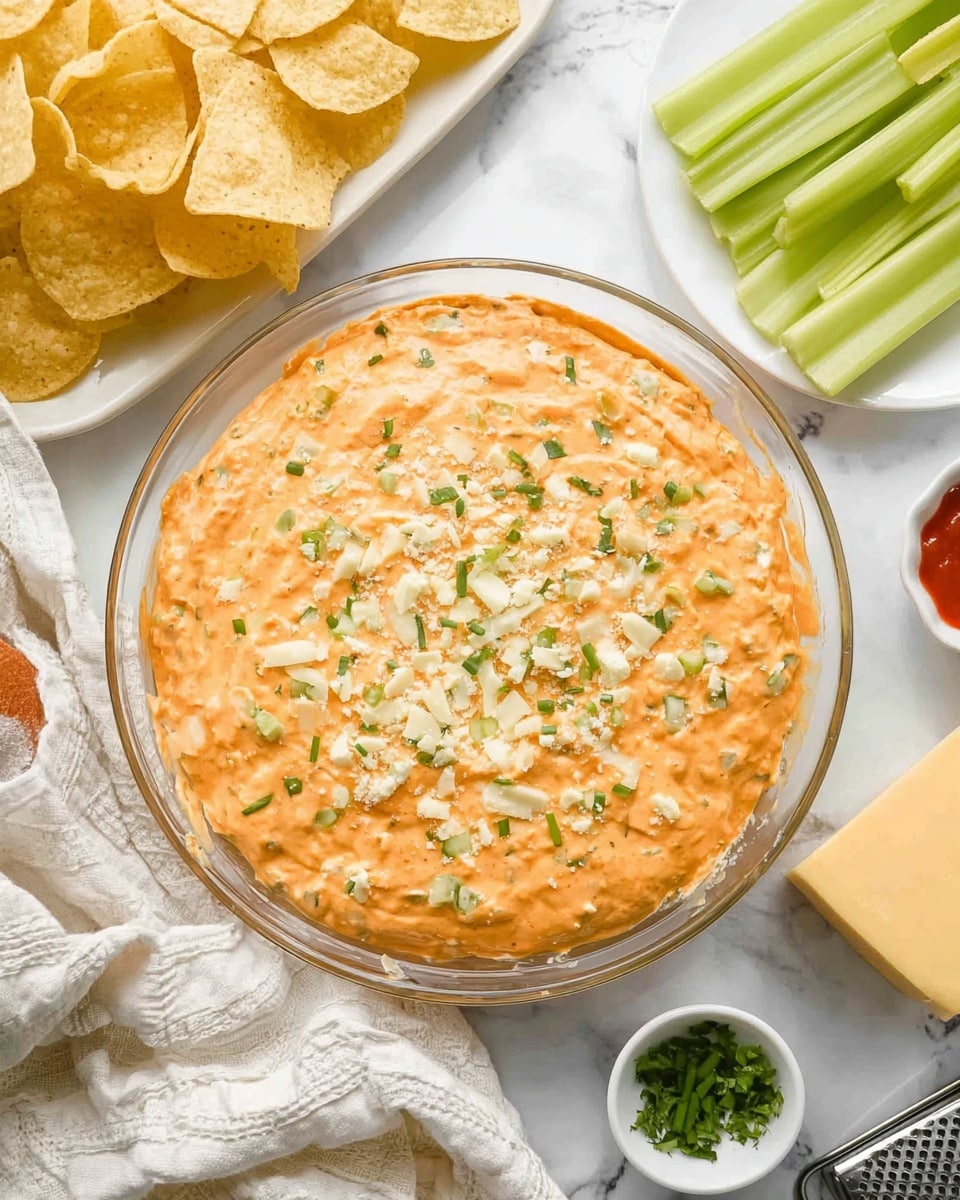 A clear glass round dish filled with one thick layer of orange creamy dip topped with small green chopped herbs and some grated white cheese scattered evenly on the surface, placed on a white marbled textured cloth background. Around the dish, there are light yellow tortilla chips stacked on the left side, several light green celery sticks on the top right, a white plate with a block of yellow cheese and a cheese grater on the bottom right, and small white bowls with a few chopped green herbs and red sauce on the left side. photo taken with an iphone --ar 4:5 --v 7
