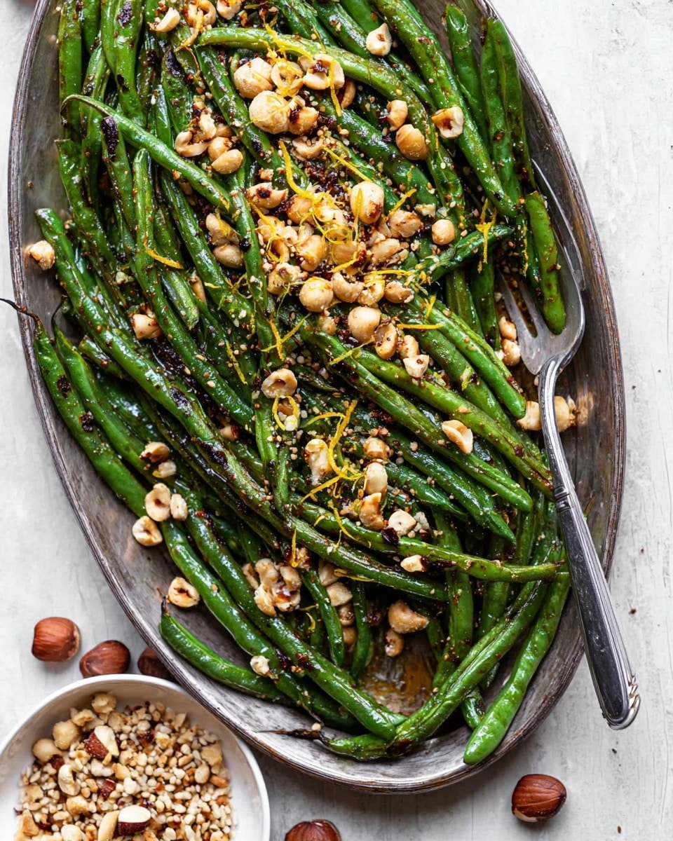 The image shows a large oval silver tray filled with vibrant green roasted green beans with charred spots, layered evenly across the tray. On top, there is a generous scattering of chopped toasted hazelnuts that add a rough texture and light brown color contrast. Thin strips of yellow zest are spread sparingly around, adding small pops of bright color. A silver fork rests on the right side of the tray. Next to the tray on a white marbled surface, a small white bowl contains extra chopped hazelnuts mixed with seeds. Whole hazelnuts are also scattered around the surface. photo taken with an iphone --ar 4:5 --v 7