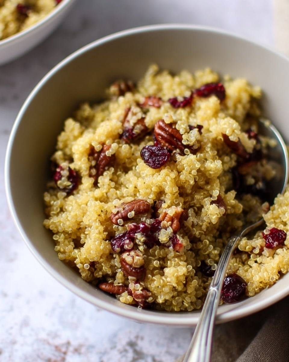 A close-up view of a bowl filled with cooked quinoa mixed with small pieces of pecans and dried cranberries. The quinoa is light yellow with a fluffy and slightly grainy texture. The pecans are brown and chunky, scattered evenly throughout, while the dried cranberries are deep red and plump. A silver spoon is partially submerged in the bowl on the right side, with its handle extending outward. The bowl is white with a smooth interior and sits on a white marbled surface. The background is softly blurred. photo taken with an iphone --ar 4:5 --v 7