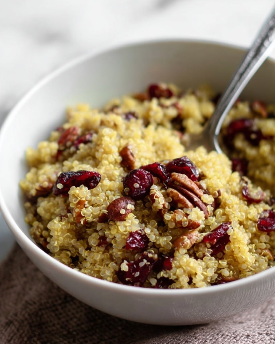 A close-up view of a bowl filled with a cooked quinoa dish mixed with small pieces of pecans and dried cranberries. The quinoa is light yellow with a fluffy texture, evenly combined with the deep red cranberries and brown nut pieces. The silver spoon rests inside the bowl on the right side, partially submerged in the quinoa mix. The bowl is white and placed on a white marbled surface. photo taken with an iphone --ar 4:5 --v 7