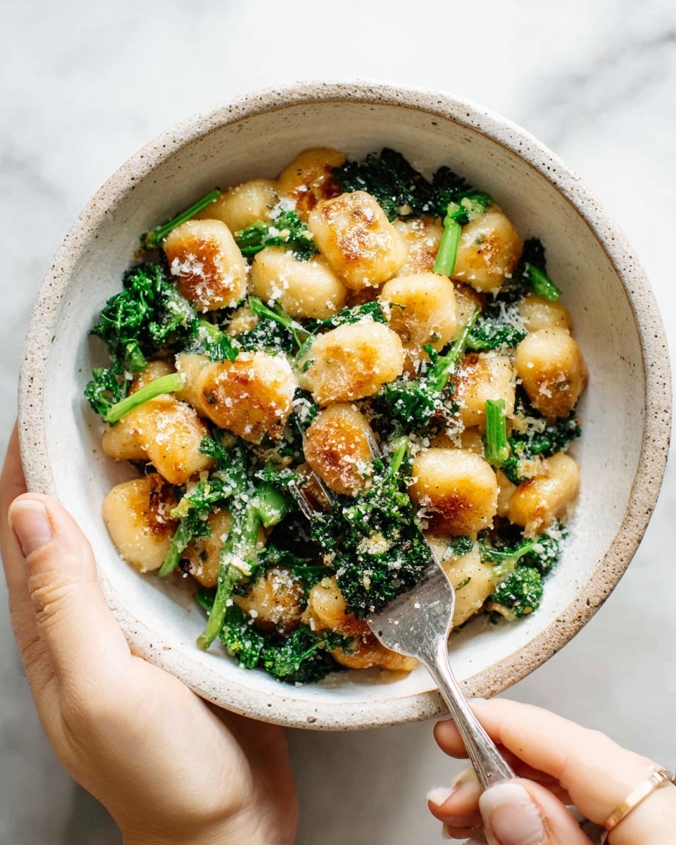 A close-up of a white speckled bowl filled with golden brown gnocchi pieces that are slightly crisp on the outside, mixed with bright green cooked broccoli rabe scattered throughout. The dish is lightly dusted with grated cheese and sits on a white marbled surface. A silver fork rests inside the bowl, and it is held by a woman's hand from below, framing the bowl in the image. photo taken with an iphone --ar 4:5 --v 7