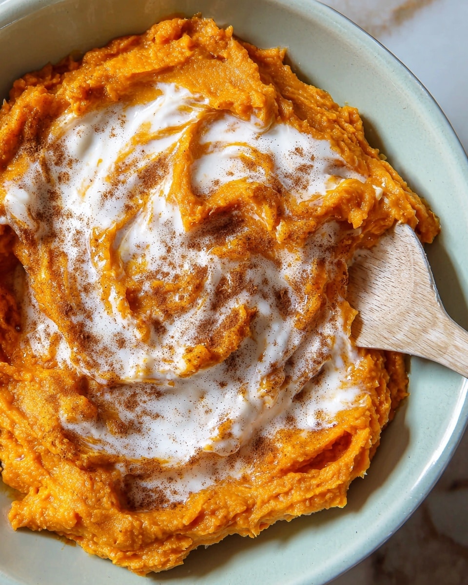 The image shows a close-up of a dish in a white bowl, featuring a mound of bright orange mashed sweet potatoes with a creamy texture. Swirled on top is a layer of white cream that lightly blends into the orange base, creating a marbled effect. The sweet potatoes are sprinkled with a dusting of dark brown cinnamon powder, adding speckled texture. On the right edge of the bowl, a light wooden spoon partially dips into the mash, touching the mixture gently. The bowl rests on a white marbled surface. photo taken with an iphone --ar 4:5 --v 7