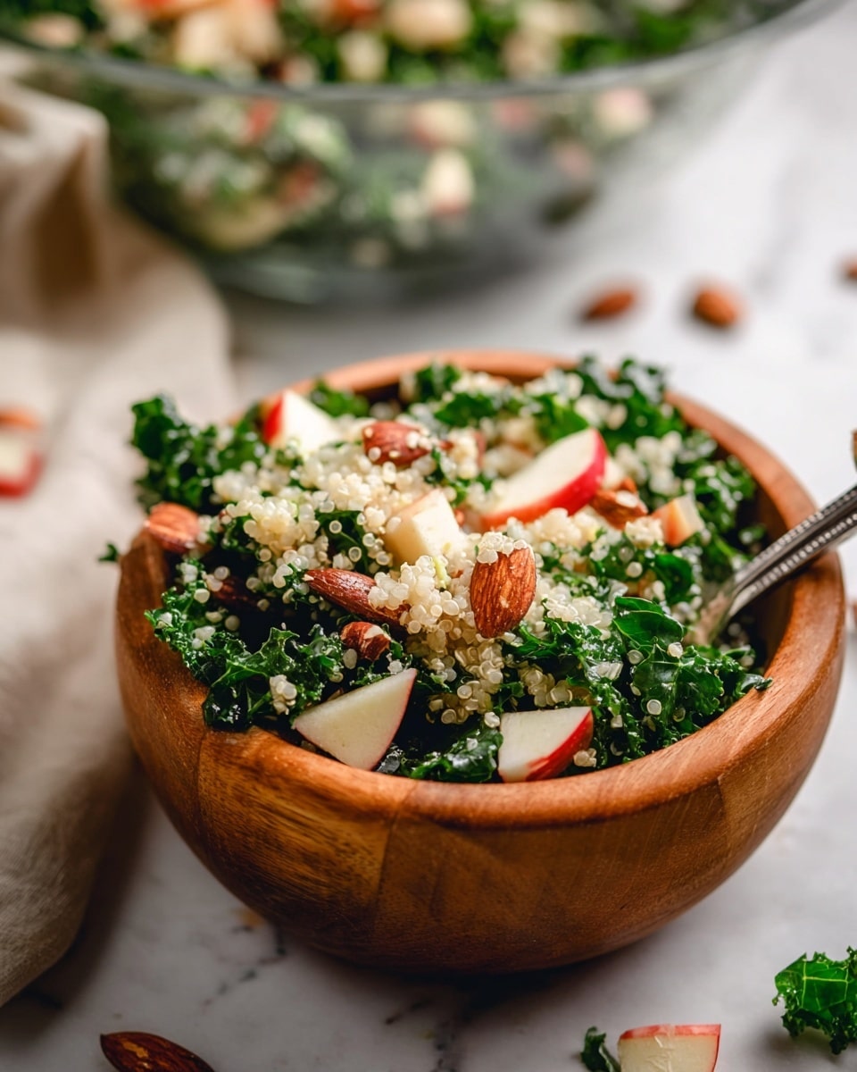 A close-up view of a wooden bowl filled with a fresh salad made of dark green kale leaves forming the base layer, topped with small white quinoa grains scattered evenly, diced red and white apple pieces adding bright color, and whole almonds giving a crunchy texture. A silver spoon is partially inside the bowl, resting on the salad. The bowl sits on a white marbled surface with some kale leaves and almonds scattered around. In the background, a clear glass bowl with more salad is slightly blurred. Photo taken with an iphone --ar 4:5 --v 7