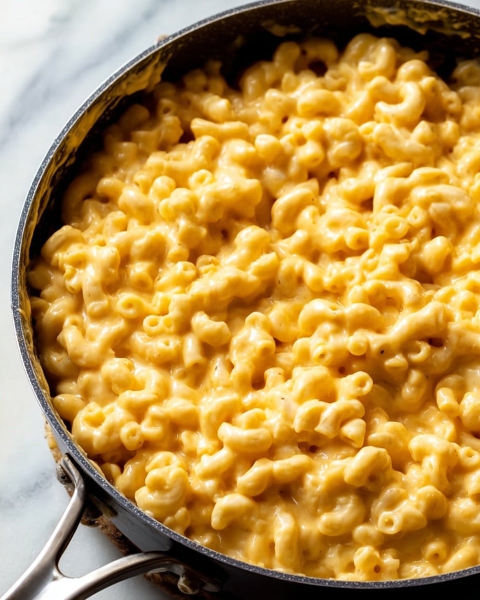 The image shows a close-up of a large pan filled with creamy macaroni and cheese. The macaroni is elbow-shaped, covered in a thick, smooth, light orange-yellow cheese sauce that looks rich and glossy. The cheese coats every piece evenly, giving the dish a soft and gooey texture. The pan is black with a shiny metal handle, set on a white marbled surface. The photo focuses on the cheesy pasta, capturing its warmth and comfort. photo taken with an iphone --ar 4:5 --v 7