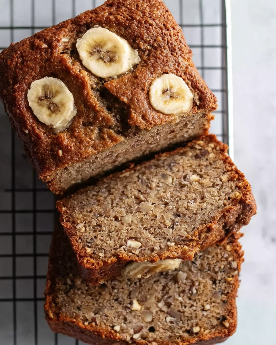 A loaf of banana bread resting on a white marbled surface, cut into three slices. The top of the loaf is golden brown with a slightly rough texture, decorated with two round banana slices embedded on the crust. The inside of the bread is dense and moist with visible small pieces of nuts scattered throughout, showing a rich brown color. The banana bread is placed on a cooling rack with a silver grid pattern, adding a subtle contrast to the warm tones of the bread. photo taken with an iphone --ar 4:5 --v 7