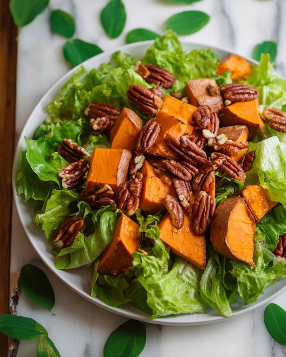 A white round plate is filled with three layers: the bottom layer is fresh green lettuce leaves with varying shades from light to medium green, providing a leafy texture. On top of the lettuce, there are chunky pieces of roasted sweet potatoes with a bright orange inner flesh and darker orange-brown skin, cut into large irregular wedges. The top layer is a generous scattering of whole pecan nuts, deep brown in color and adding a textured, slightly glossy finish to the dish. The background is a white marbled texture with scattered green leaves, and a woman's hand is not visible in this image. photo taken with an iphone --ar 4:5 --v 7