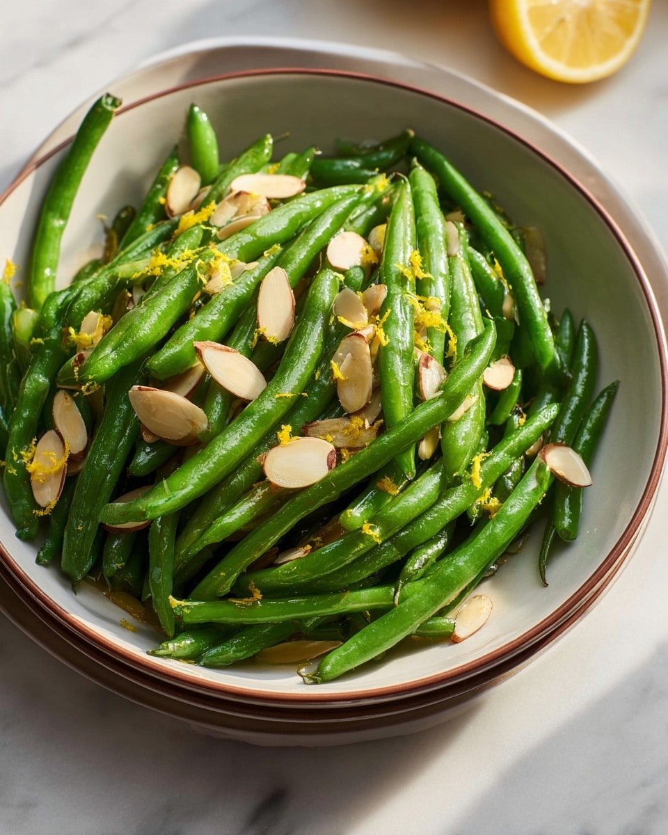 A bowl filled with bright green cooked green beans lies on a white marbled surface, topped with thin, light brown almond slices scattered over the beans. Some small bits of yellow lemon zest are sprinkled on top, adding a touch of color contrast. The bowl is white with a light brown rim and has a smooth, slightly textured surface. In the background, there is a glass with a golden drink that catches the light. Photo taken with an iphone --ar 4:5 --v 7