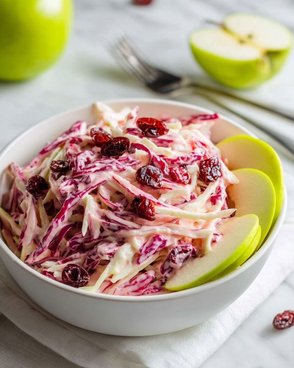 A white bowl filled with a creamy coleslaw salad made of thin white and deep pink cabbage strands mixed with a pale pink dressing. The dish is topped with dark red dried cranberries scattered across the surface. Fresh green apple slices with white inner flesh are placed on top and around the coleslaw, adding fresh color contrast. In the background, a halved green apple and a white napkin with a silver fork rest on a white marbled surface. Photo taken with an iphone --ar 4:5 --v 7