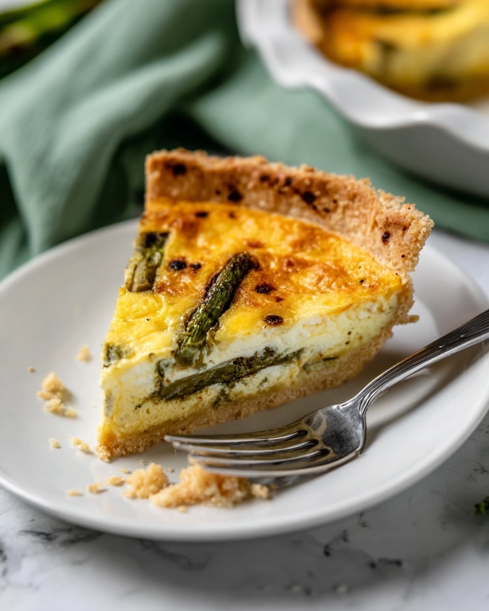 A single slice of quiche sits on a white plate, showing three main layers: the top layer is golden yellow and slightly browned with small black specks, the middle layer is creamy white with visible pieces of green asparagus, and the bottom layer is a thicker pale yellow custard resting on a light brown, crumbly crust with a raised edge. The crust looks flaky and firm. A silver fork lies next to the slice on the plate, and crumbs are scattered near the slice. The background shows a soft green cloth and another part of the quiche in a white dish on a white marbled surface. Photo taken with an iphone --ar 4:5 --v 7