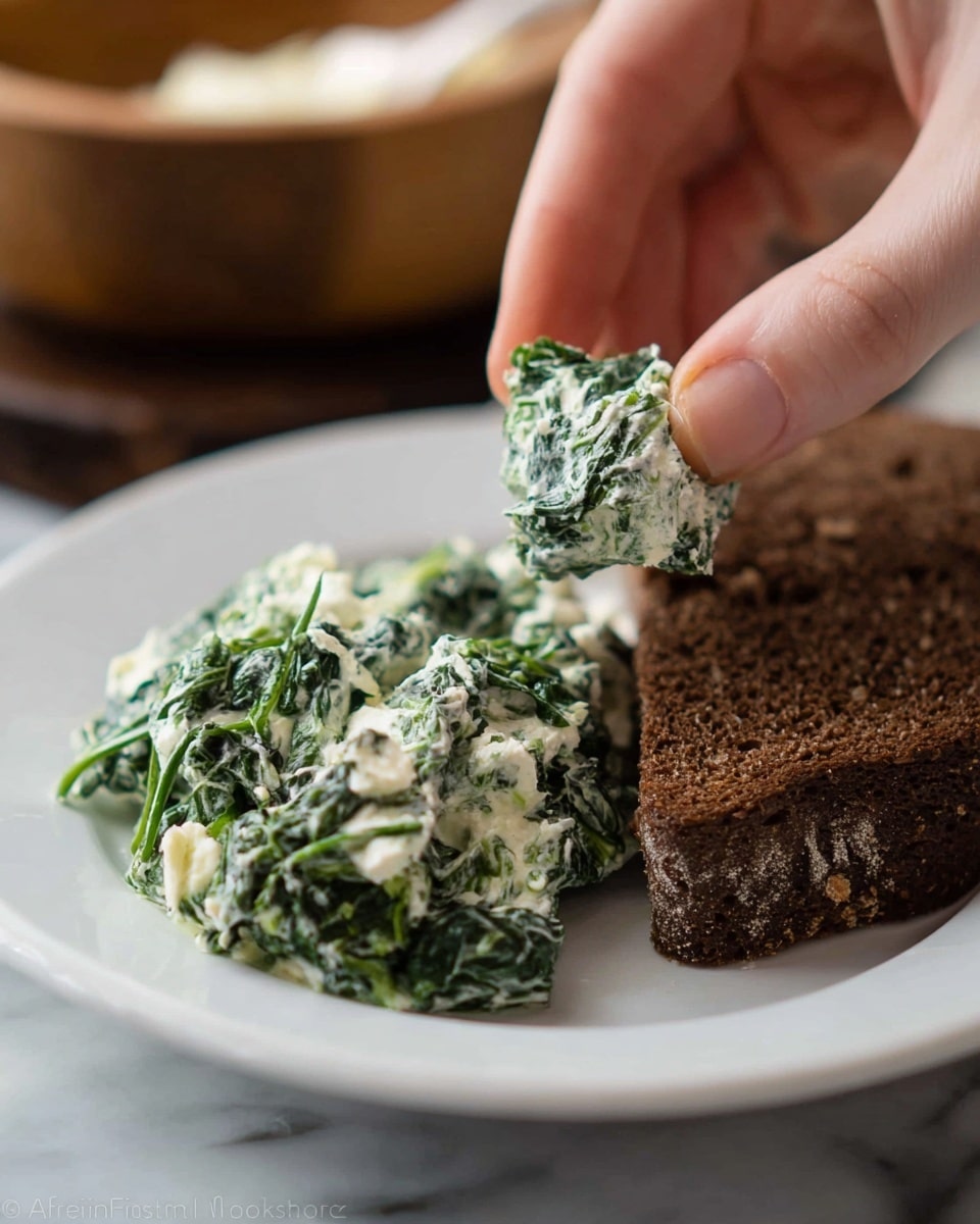 A white plate holds a serving of green spinach mixed with creamy white cheese or sauce, spread loosely on one side. Next to the mixture, there is a thick slice of dark brown bread with a rough texture. A woman’s hand is pinching a small piece of the dark brown bread, scooping the spinach and cheese mix. The background is softly blurred showing a wooden bowl. The plate is placed on a white marbled surface. photo taken with an iphone --ar 4:5 --v 7