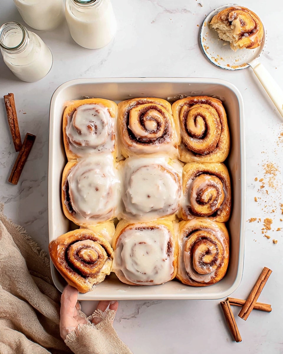 The image shows nine cinnamon rolls arranged in a white rectangular baking dish, with one roll missing from the center, leaving an empty space. Each cinnamon roll has a golden-brown, slightly glossy baked outer layer with darker brown swirls of cinnamon visible in the spiral on top. Six of the rolls are topped with a thick, creamy white icing that has a smooth texture and small visible specks, while three rolls remain without icing, showing their brown cinnamon spirals clearly. The dish is on a white marbled surface with two cinnamon sticks placed to the lower left. A woman's hand holds one cinnamon roll piece just outside the dish on the left side. There are two glass bottles, likely with milk or cream, near the top left corner, and a beige cloth rests to the right side of the dish. In the top right corner, there is a small white spatula resting on a small tart pan with crumbs around. photo taken with an iphone --ar 4:5 --v 7