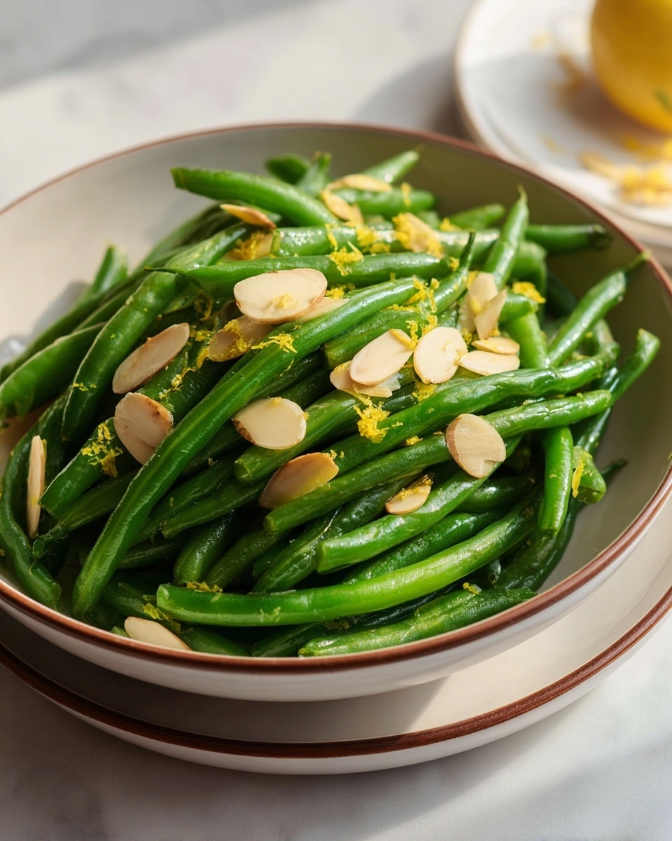 A bowl filled with bright green cooked green beans forms the base layer, with a glossy texture showing they are lightly coated in oil or butter. Scattered on top are thinly sliced almond pieces, light brown on one side and cream on the other, adding a crunchy contrast. Small bits of yellow lemon zest are sprinkled across the green beans and almonds, giving a fresh and colorful touch. The bowl is white with a subtle brown rim and rests on a white marbled surface, with soft natural light casting gentle shadows. photo taken with an iphone --ar 4:5 --v 7