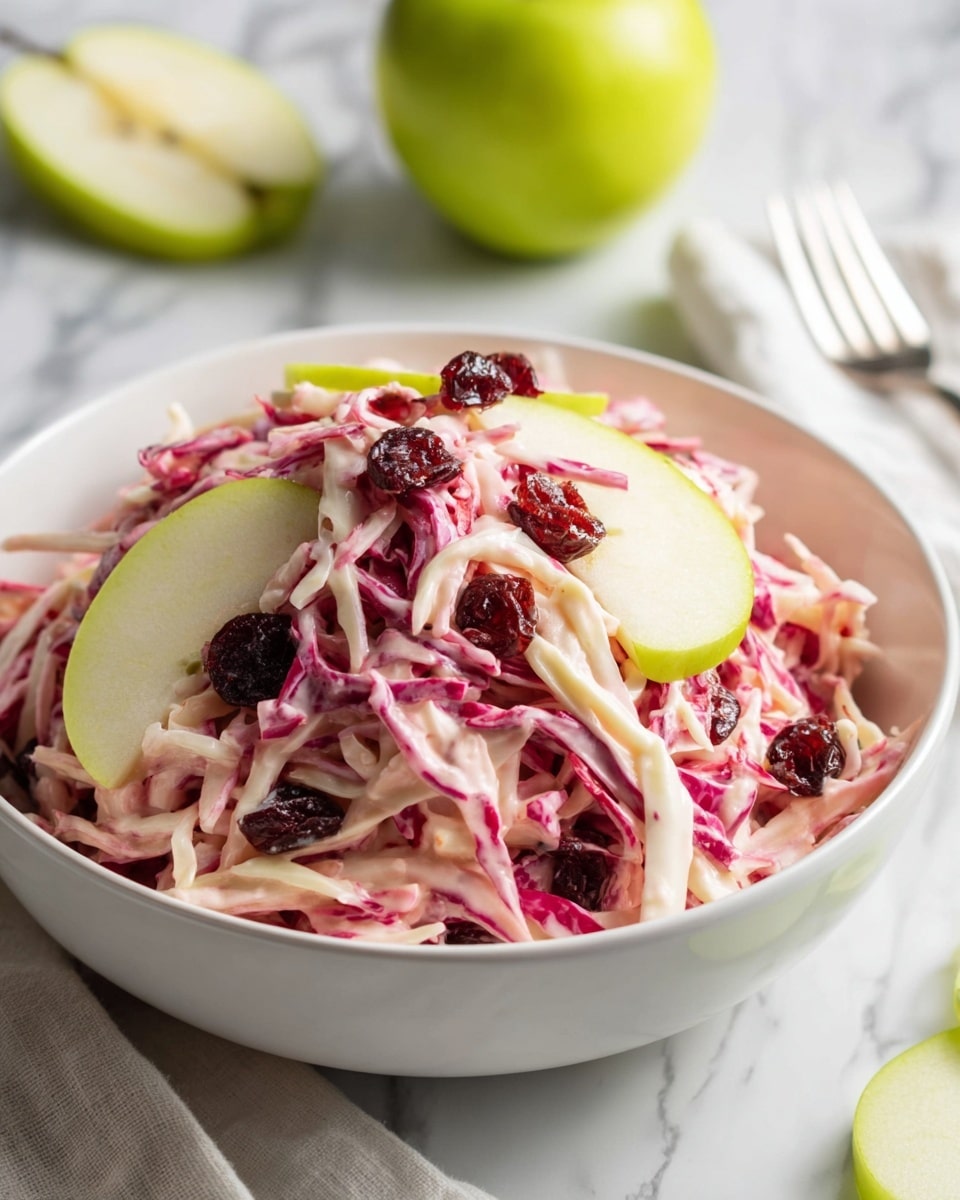 A white bowl filled with a creamy red cabbage slaw, showing thin pink and white cabbage strips coated in a light pink dressing. Scattered on top are dark red dried cranberries, and fresh green apple slices with pale flesh are arranged around and on the slaw, adding color contrast. The bowl sits on a white marbled surface with a white cloth near a silver fork and a green apple cut in half nearby. photo taken with an iphone --ar 4:5 --v 7