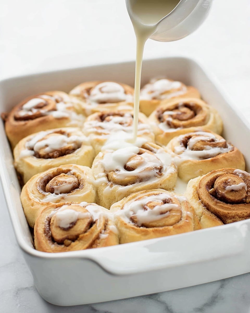 A soft, light brown cinnamon roll with a gooey glaze on top sits on a white plate. The roll features a spiral shape with layers visible, showing a moist and fluffy interior texture. A section of the roll has been torn off, revealing the airy, slightly crumbly inside with some sticky glaze spreading out on the plate. There are a few scattered crumbs around the base. The background and surface have a white marbled texture, creating a clean and simple look. Photo taken with an iphone --ar 4:5 --v 7