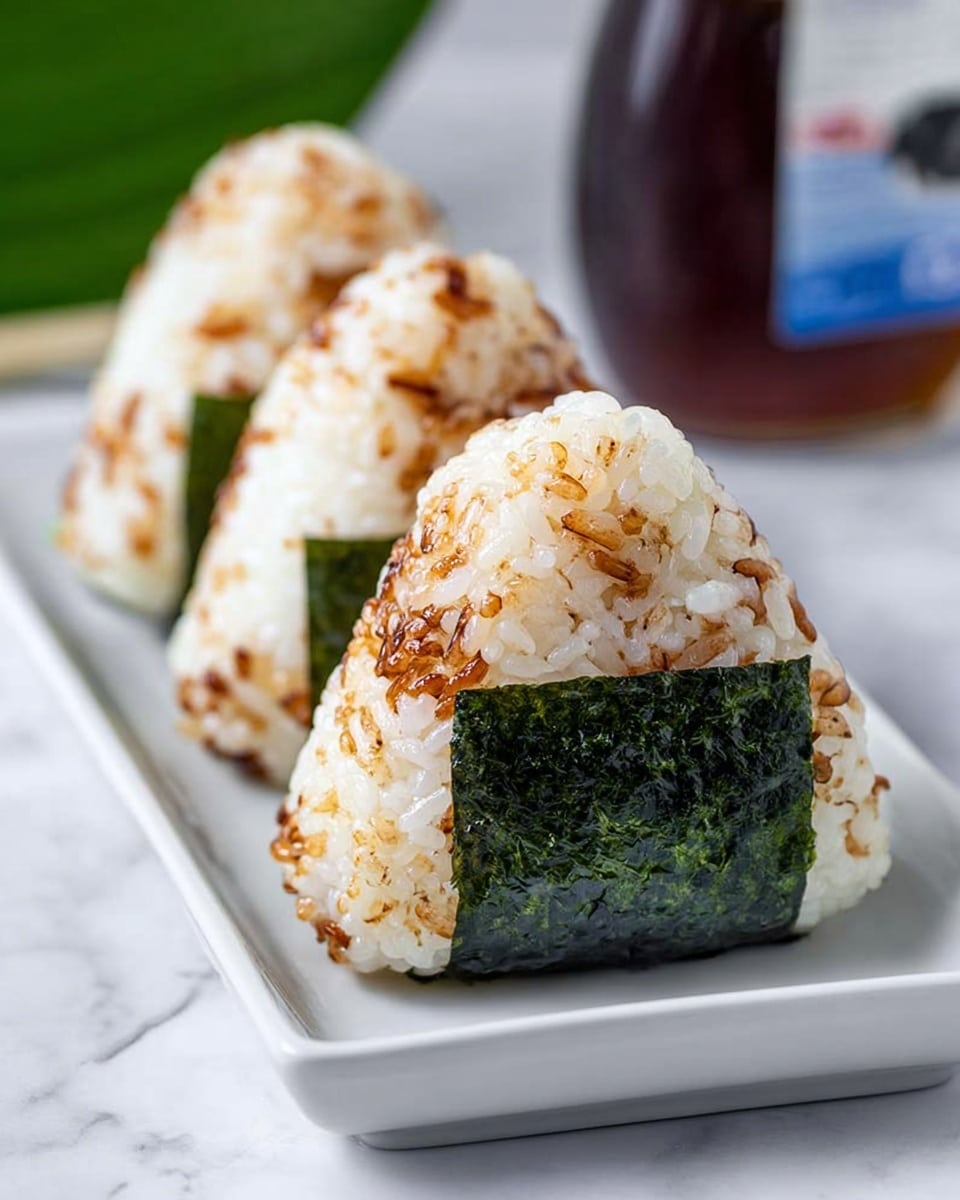 Three triangular rice balls with toasted brown spots mixed in the white rice form the main layers, each wrapped at the base with a glossy dark green seaweed strip. They are placed in a line on a long white rectangular plate resting on a white marbled surface, with a blurred bottle and green leaf in the background. The close-up view shows the rice grains’ sticky texture clearly. Photo taken with an iphone --ar 4:5 --v 7