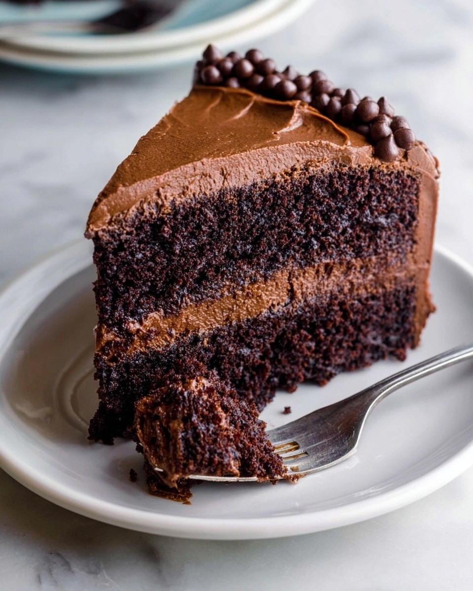 A slice of two-layer dark chocolate cake sits on a white plate, with a thick middle layer of smooth chocolate frosting. The top layer is covered with a smooth, slightly textured frosting and decorated with small, shiny chocolate drops along the edge. The cake layers look moist and dense, with a rich chocolate color. A piece of cake with frosting is held by a fork resting on the plate in front of the slice. The surface beneath the plate is a white marbled texture. photo taken with an iphone --ar 4:5 --v 7