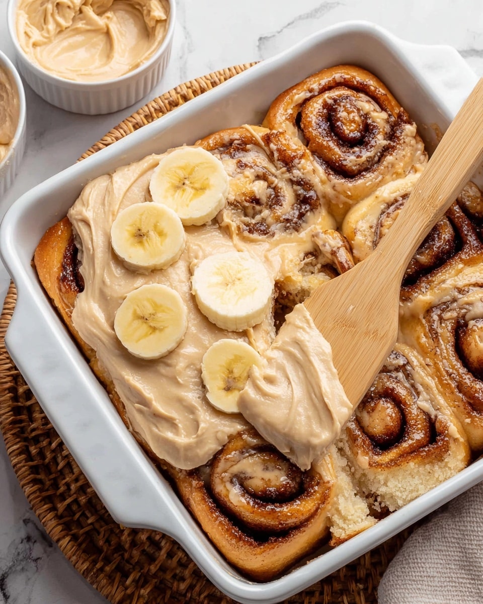 A white rectangular baking dish with a layer of six cinnamon rolls inside, showing a golden brown color with dark brown swirls of cinnamon and a soft, fluffy texture. Part of the cinnamon rolls is covered with a creamy beige frosting, which is being spread by a wooden spatula. On top of the frosting, three slices of fresh banana are placed, adding a light yellow accent. To the side, a small white bowl holds more beige frosting. The dish is set on a woven basket tray, all placed on a white marbled surface. photo taken with an iphone --ar 4:5 --v 7