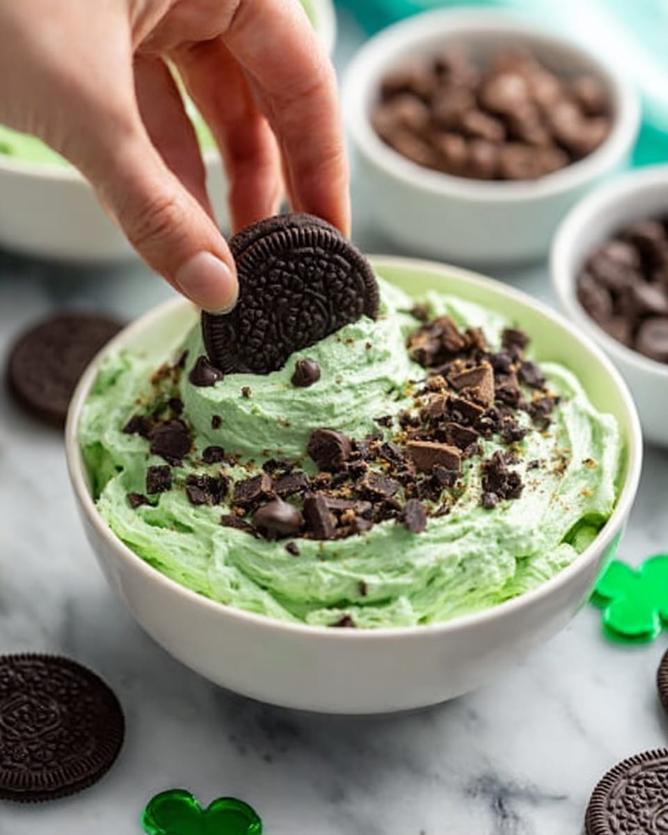A white bowl filled with a thick, green mint-flavored mixture topped with small chocolate chips, chopped dark chocolate pieces, and a whole chocolate cookie being placed into the mixture by a woman's hand from the top left. The green layer has a creamy, slightly fluffy texture, with the toppings resting unevenly on top. In the background, there are white bowls containing more chocolate cookies and chocolate chips on a white marbled surface. Photo taken with an iphone --ar 4:5 --v 7