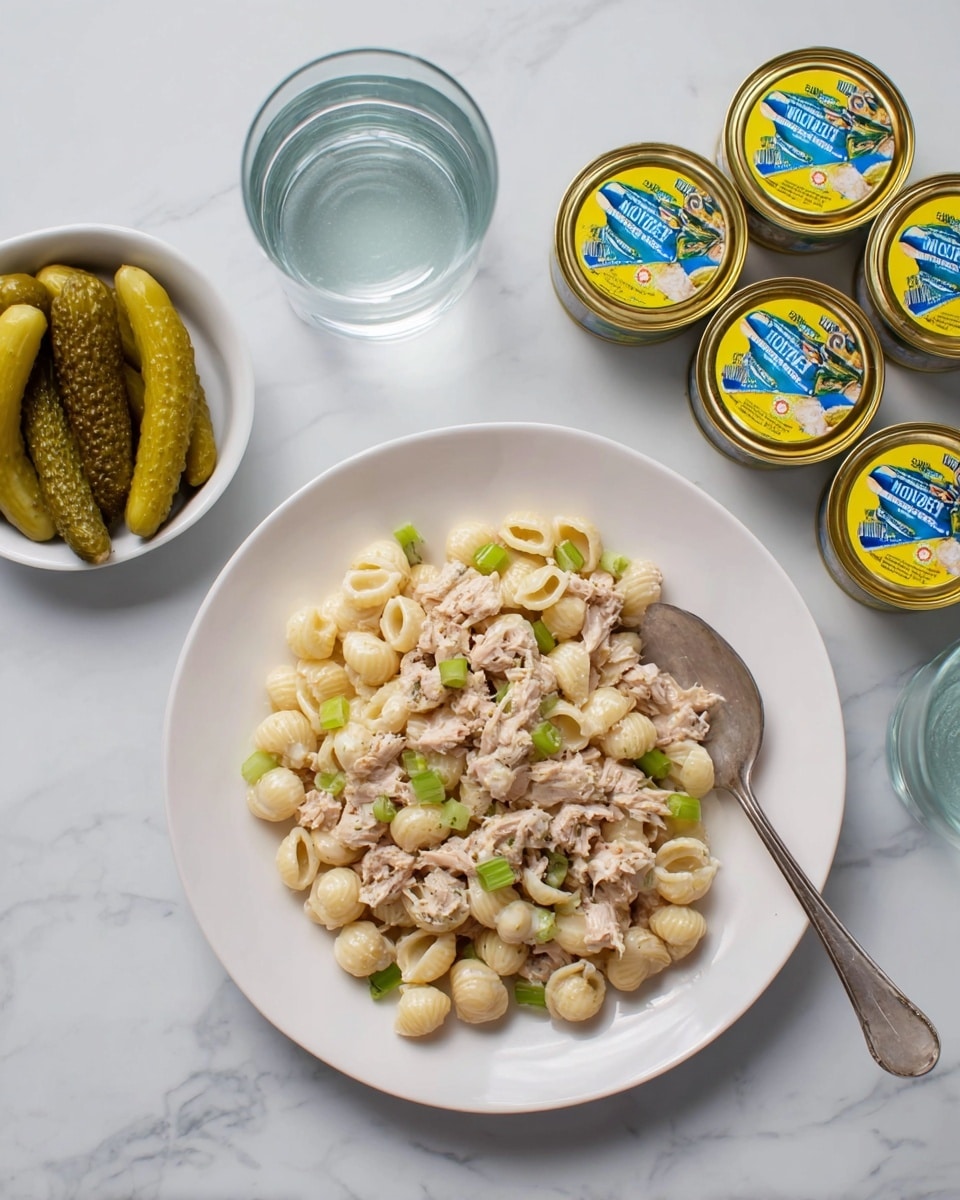 The image shows a white plate filled with a tuna pasta salad made of small shell pasta mixed with light pink tuna flakes and small pieces of green celery, creating a mix of creamy beige and fresh green colors with a slightly creamy texture. A silver spoon is placed on the right side of the plate partly resting in the salad. Around the plate, there are three closed cans of tuna with bright yellow and blue labels. To the left of the plate, a small white bowl contains four sliced pickle spears of a pale green color, and above are two clear glasses of water with visible reflections. The whole scene is set on a white marbled surface. photo taken with an iphone --ar 4:5 --v 7