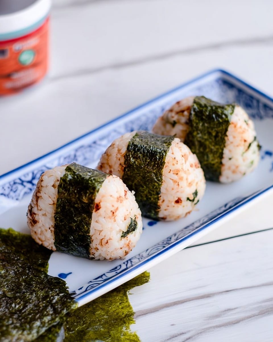 The image shows three oval-shaped rice balls with a light brown seasoning mixed in, placed side by side on a white rectangular plate with blue patterns along the edges. Each rice ball has a thin strip of dark green seaweed wrapped around its bottom. The plate sits on a white marbled surface, with a large sheet of green seaweed partially visible next to it. A blurred container with a red label is in the background. photo taken with an iphone --ar 4:5 --v 7