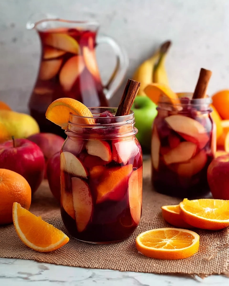 A clear glass jar filled with a deep red drink showing visible layers of orange slices, red berries, and apple slices beneath the surface, topped with a whole cinnamon stick and garnished with a small orange wedge near the rim; in the background, there is another similar jar and a clear glass pitcher filled with more of the red drink containing floating fruit slices and cinnamon sticks; the jars and pitcher sit on a brown cloth resting on a white marbled surface, surrounded by halved bright orange oranges and sliced red apples, with a few whole apples and bananas blurred in the background, creating a rich, fresh, and colorful fruit drink setting. photo taken with an iphone --ar 4:5 --v 7