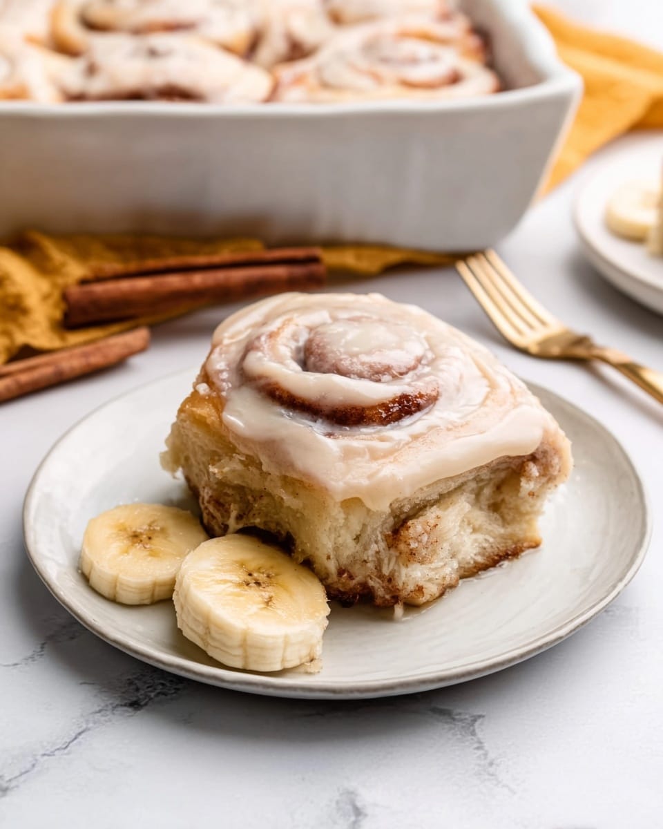 A single cinnamon roll sits on a small white plate with a thick layer of smooth, light cream cheese frosting covering the top and slightly dripping down the sides. The roll is golden brown with visible swirls of cinnamon filling, soft and fluffy in texture, and slightly glazed. Next to the cinnamon roll, two round banana slices rest on the plate revealing a soft, pale yellow inside. The plate is set on a white marbled surface with a gold fork nearby and a woman’s hand gently resting close to the plate. In the background, there is a white baking dish filled with more cinnamon rolls glazed similarly, with a cinnamon stick placed beside it. photo taken with an iphone --ar 4:5 --v 7
