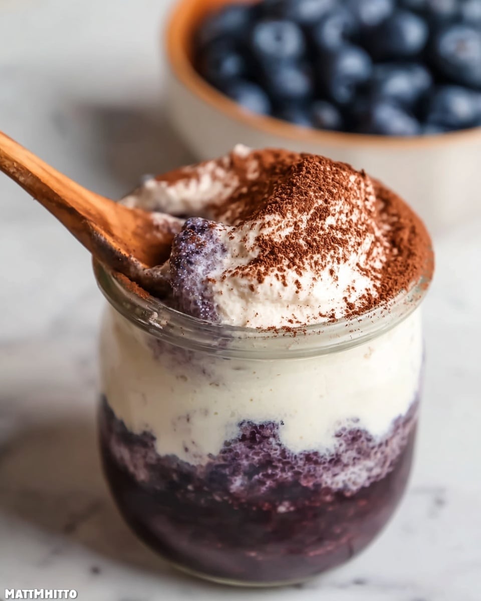 The image shows a clear glass jar filled with three visible layers: the bottom layer is a dark purple textured mixture, the middle layer is creamy white with purple streaks blending into it, and the top layer is thick white cream sprinkled with a light dusting of brown powder. A wooden spoon is placed inside the jar, partially resting on the creamy top layer. The jar sits on a wooden board with some powder sprinkled around its base, and the background is a white marbled texture. Photo taken with an iphone --ar 4:5 --v 7