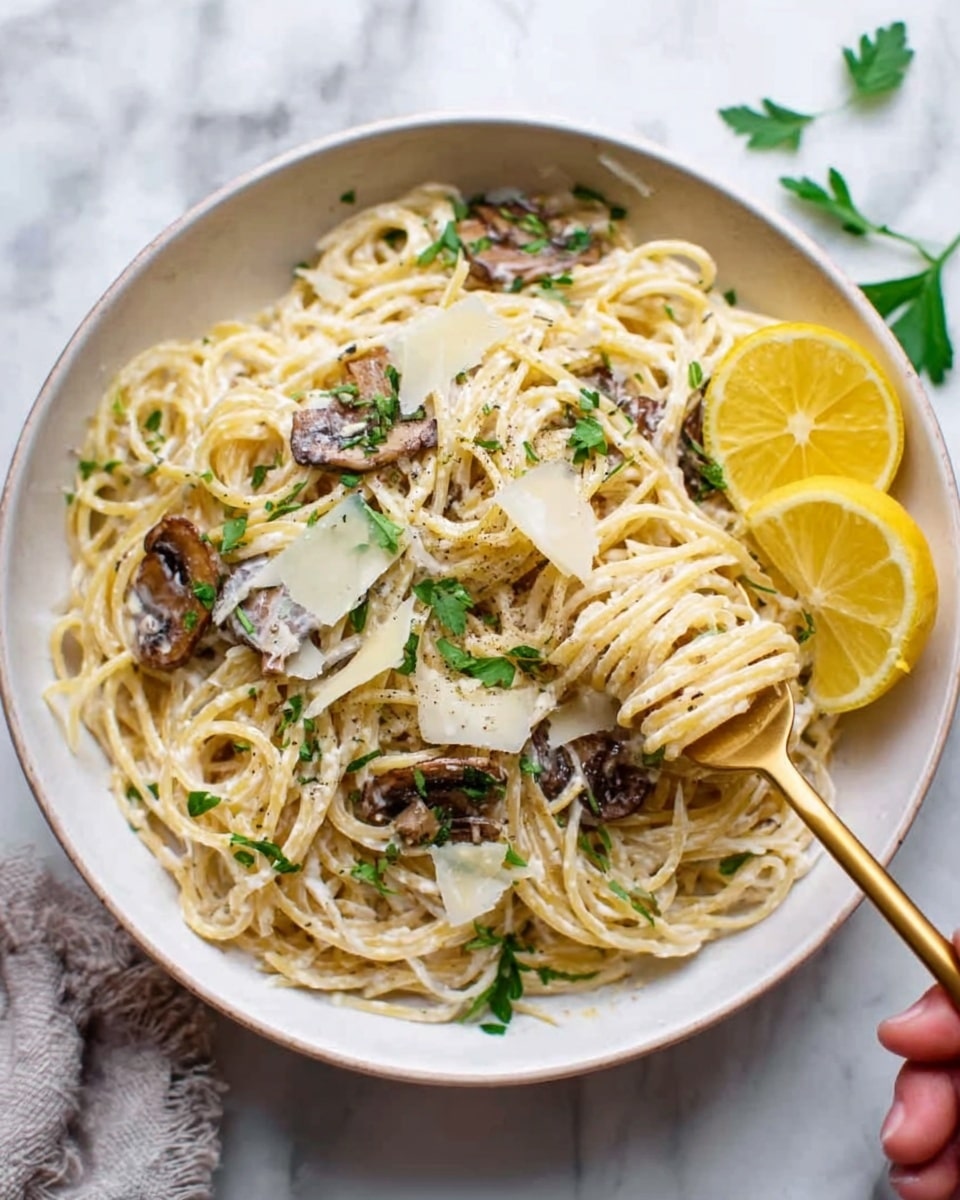 A white bowl filled with a serving of spaghetti pasta, layered with creamy white sauce and topped with browned slices of mushrooms. Thin shavings of pale yellow cheese are scattered over the top, along with small green parsley leaves adding fresh color. Two bright yellow lemon wedges rest on the right side of the bowl. A woman's hand holds a gold fork, gathered with some pasta and sauce, lifting it above the bowl. The whole scene is set on a white marbled surface. Photo taken with an iphone --ar 4:5 --v 7