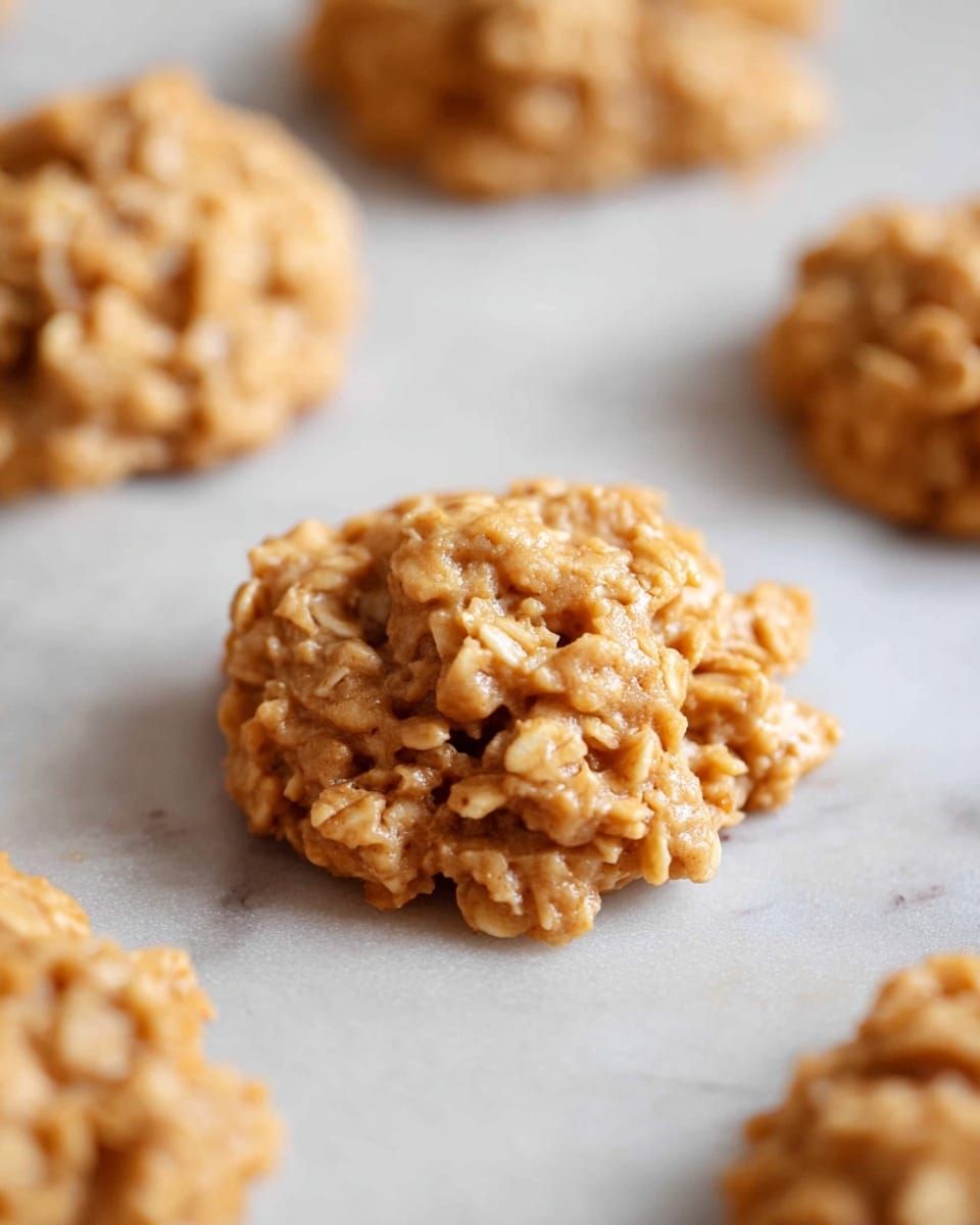 The image shows a close-up of several small, rough-textured cookies placed on a white marbled surface. The cookies are light brown with a lumpy, uneven shape, indicating oats or similar grains mixed into the dough. Each cookie has a thick, chunky look with visible pieces throughout, and they are spaced out on the surface. The focus is on one cookie in the center, which is in sharp detail, while the others in the background are softly blurred. photo taken with an iphone --ar 4:5 --v 7