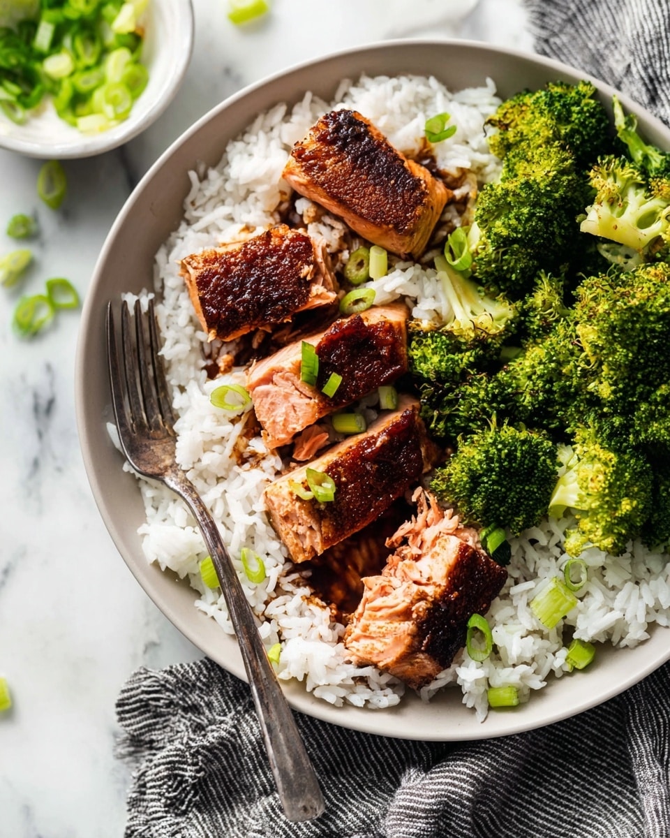 A white round plate holds a serving of white rice as the base layer, with some soy sauce or dark sauce mixed lightly through it. On top of the rice, there are five pieces of grilled salmon that show a crispy, dark brown crust on the outside and soft pink inside. The salmon pieces are styled in the center of the plate. To the right side, bright green steamed broccoli florets lay on the rice, adding a fresh and vibrant color contrast. Small slices of green onion are scattered over the salmon and rice, adding texture and a pop of light green. A metal fork rests on the left side of the plate on a gray striped cloth, and a small white bowl with more green onion slices sits blurred in the top left corner, all placed on a white marbled surface. photo taken with an iphone --ar 4:5 --v 7