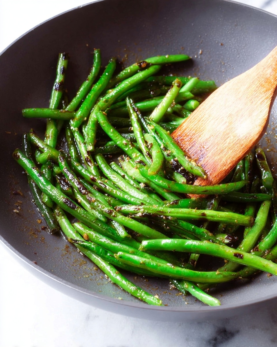 The image shows a pan filled with cooked green beans that have some charred black spots, giving them a slightly burnt texture. The green beans are bright green and appear fresh and shiny with oil coating them. A wooden spatula is partially stirring the green beans from the right side, and the pan has a dark grey, smooth surface. The background is a white marbled texture. photo taken with an iphone --ar 4:5 --v 7