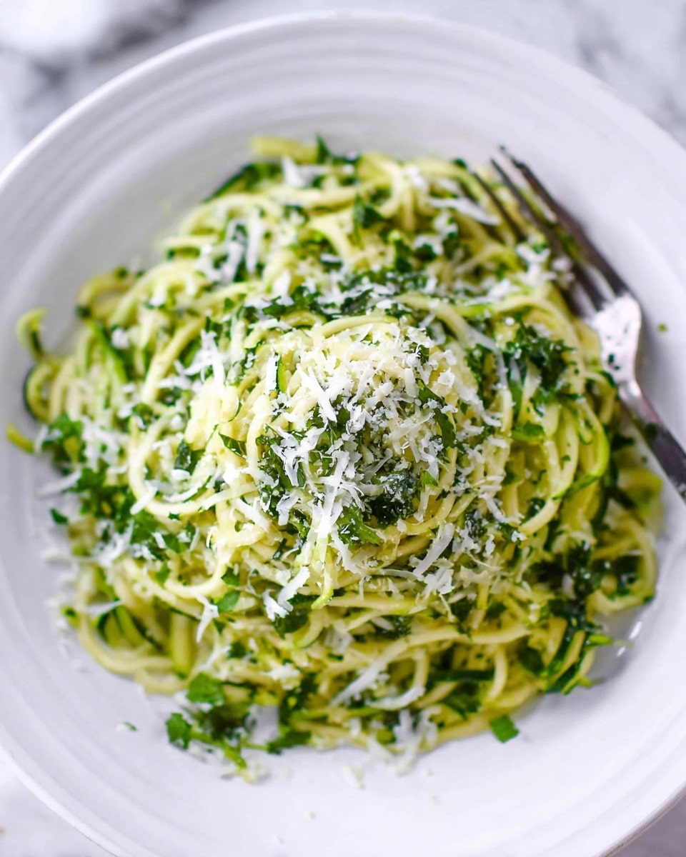 A white plate shows a mound of thin spiralized zucchini noodles colored pale green in the base layer, mixed with darker green leafy herbs scattered throughout. On top, there is a fine sprinkle of white grated cheese that adds a light dusting effect across the noodles. The texture is soft with some visible small pieces of green herbs and cheese bits, creating a fresh and light look. A fork rests on the side of the plate against a white marbled surface. Photo taken with an iphone --ar 4:5 --v 7