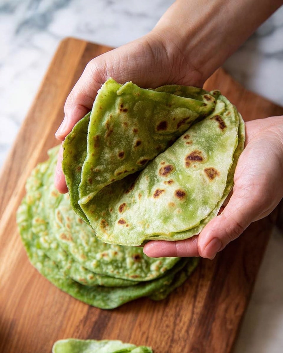 A pair of woman's hands holding a folded green flatbread, showing a soft, textured inside and a lightly bubbled outside with small brown spots. Underneath, more green flatbreads lie flat on a wooden board. The background is a white marbled texture. photo taken with an iphone --ar 4:5 --v 7