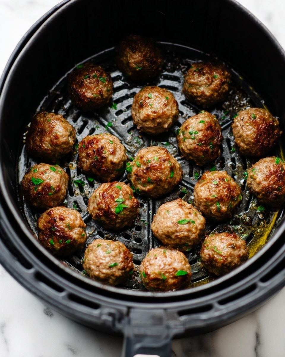 A black air fryer basket filled with 18 browned meatballs, each one round and textured with a slight crisp on the outside. Small bits of green herbs are sprinkled lightly over the meatballs, adding a fresh touch of color. The basket's perforated bottom is visible beneath the meatballs, showing some oil glistening around them. The background is a white marbled texture. photo taken with an iphone --ar 4:5 --v 7