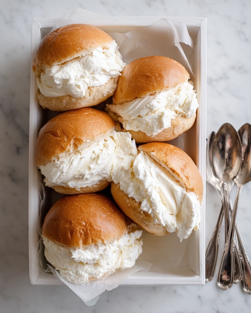 The image shows five soft, golden-brown buns filled generously with thick white cream, arranged closely inside a white square box. Each bun's top half is pulled apart slightly to hold an ample layer of smooth, fluffy cream that almost spills out, creating a puffy and inviting look. The box sits on a white marbled surface, to the right of it are two metal spoons placed side by side, and the overall scene conveys a fresh and homemade feel. photo taken with an iphone --ar 4:5 --v 7