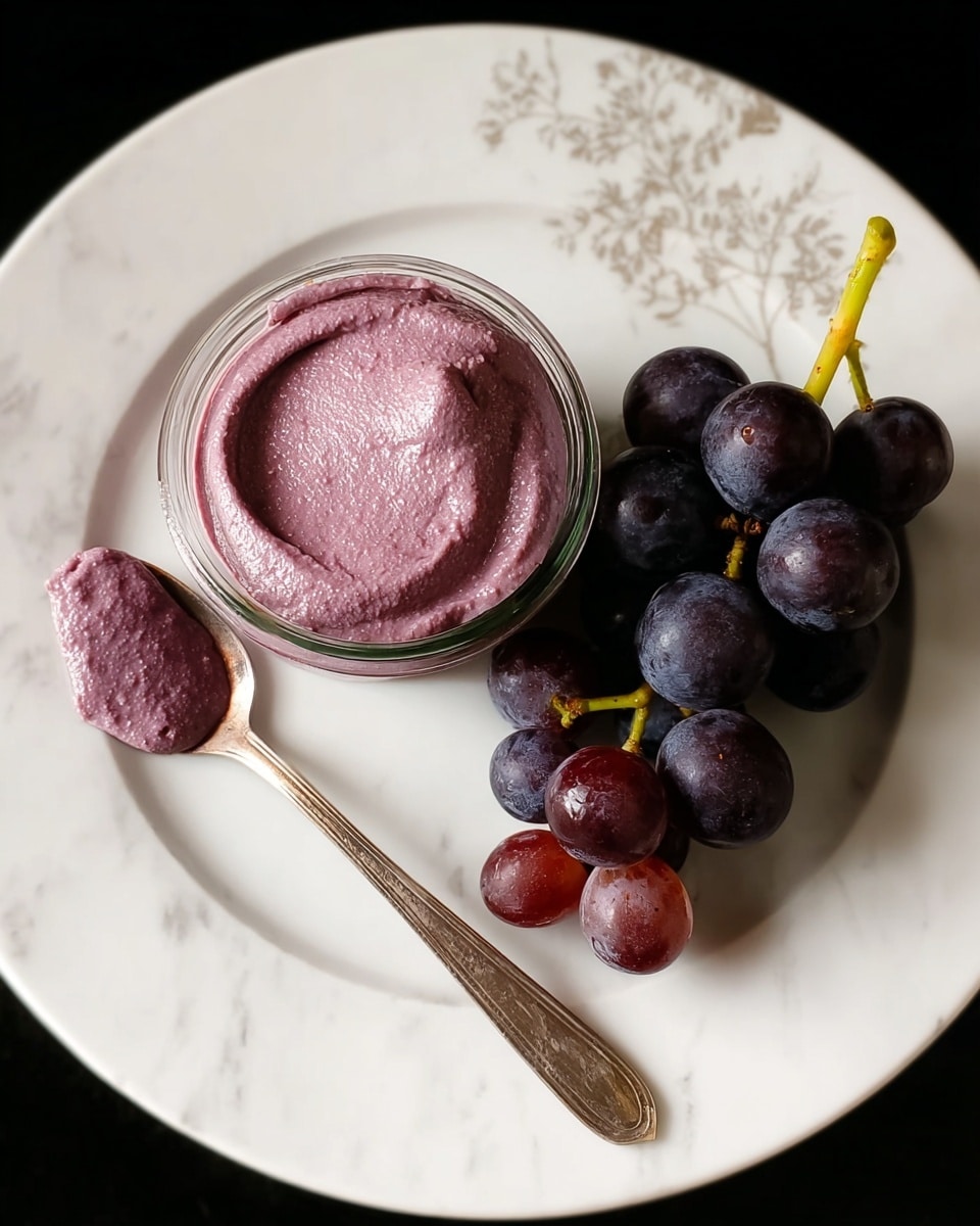A white plate with a subtle floral pattern holds a small glass jar filled with a thick, creamy purple spread that has a smooth, slightly textured surface. Next to the jar, a cluster of dark purple grapes with a light green stem rests on the right side of the plate. On the left side, a silver spoon lies on the plate, with a dollop of the same purple spread on it. The background is a white marbled texture. Photo taken with an iphone --ar 4:5 --v 7