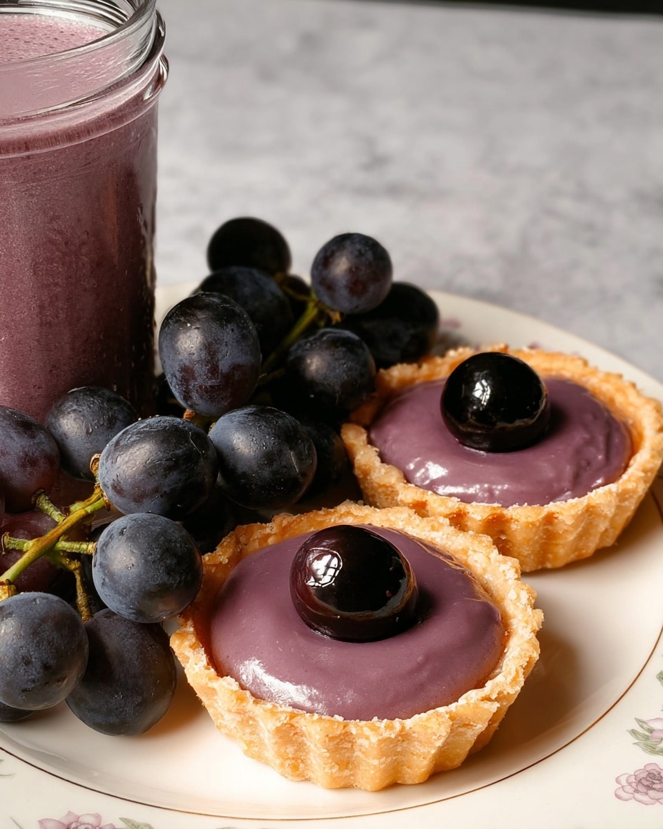 Two small tarts sit side by side on a white plate with a subtle floral pattern, placed on a white marbled background. Each tart has a golden, flaky crust with a glossy purple filling that looks smooth and creamy, topped with a single shiny black grape in the center. Next to the tarts, a small bunch of dark purple grapes with green stems rests on the plate, adding a fresh and natural touch. A glass container filled with purple smoothie or pudding is partially visible on the left side of the image. Photo taken with an iphone --ar 4:5 --v 7