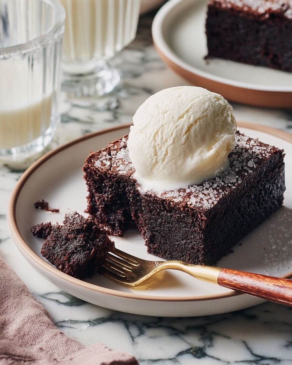 A thick, dark chocolate cake square sits on a white plate with a light brown rim. The top of the cake is textured with coarse sugar crystals and is crowned by a large dollop of smooth, creamy white ice cream. Next to the cake, a piece has been cut out, showing the moist and soft crumb inside. A fork with a wooden handle rests on the plate, with the cut piece of cake on it. The plate is set on a white marbled surface with a faint grey pattern, and there is a glass of milk and another plate with chocolate cake blurred in the background. photo taken with an iphone --ar 4:5 --v 7