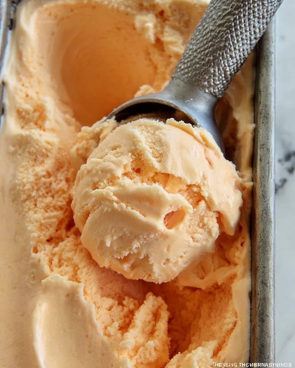 The image shows a close-up of a metal container filled with creamy, pale orange ice cream that has a smooth and slightly chunky texture. A metal ice cream scooper rests inside, scooping a soft mound of the ice cream with visible folds and gentle curves. The metal container has a patterned surface inside, and the ice cream looks freshly scooped with a shiny, moist finish. The background is a white marbled texture. photo taken with an iphone --ar 4:5 --v 7